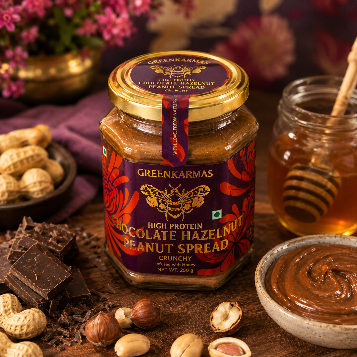 A glass jar of Greenkarmas High Protein Chocolate Hazelnut Peanut Spread sits on a wooden table surrounded by peanuts, chocolate pieces, hazelnuts, a bowl of spread, and a honey dipper. Another jar and blurred flowers appear in the background.