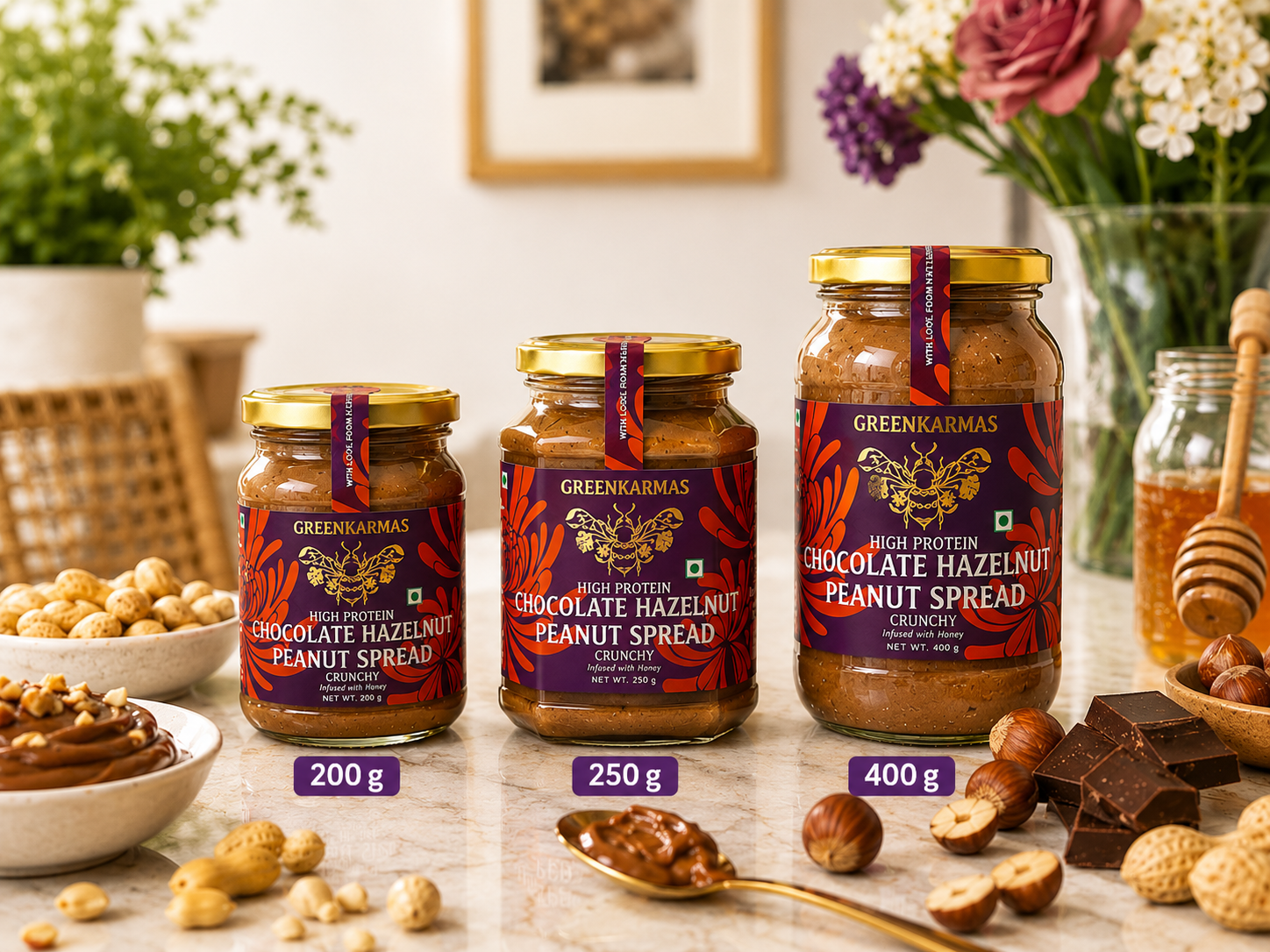 Three jars of GreenKarmas Chocolate Hazelnut Peanut Spread in 200g, 250g, and 400g sizes, arranged on a marble countertop with peanuts, chocolate pieces, and a honey pot in a warm kitchen setting.
