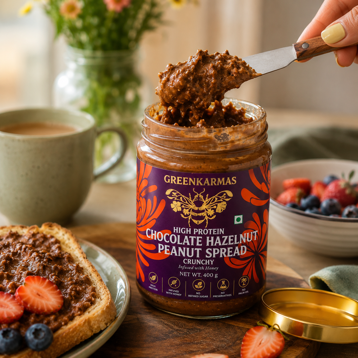 A jar of GREENKARMAS Chocolate Hazelnut Peanut Spread sits on a wooden table with a hand holding a knife above it, spreading the thick brown spread. The scene includes a slice of toast with strawberries, a cup of coffee, and a bowl of fresh berries in a warm, styled kitchen setting.