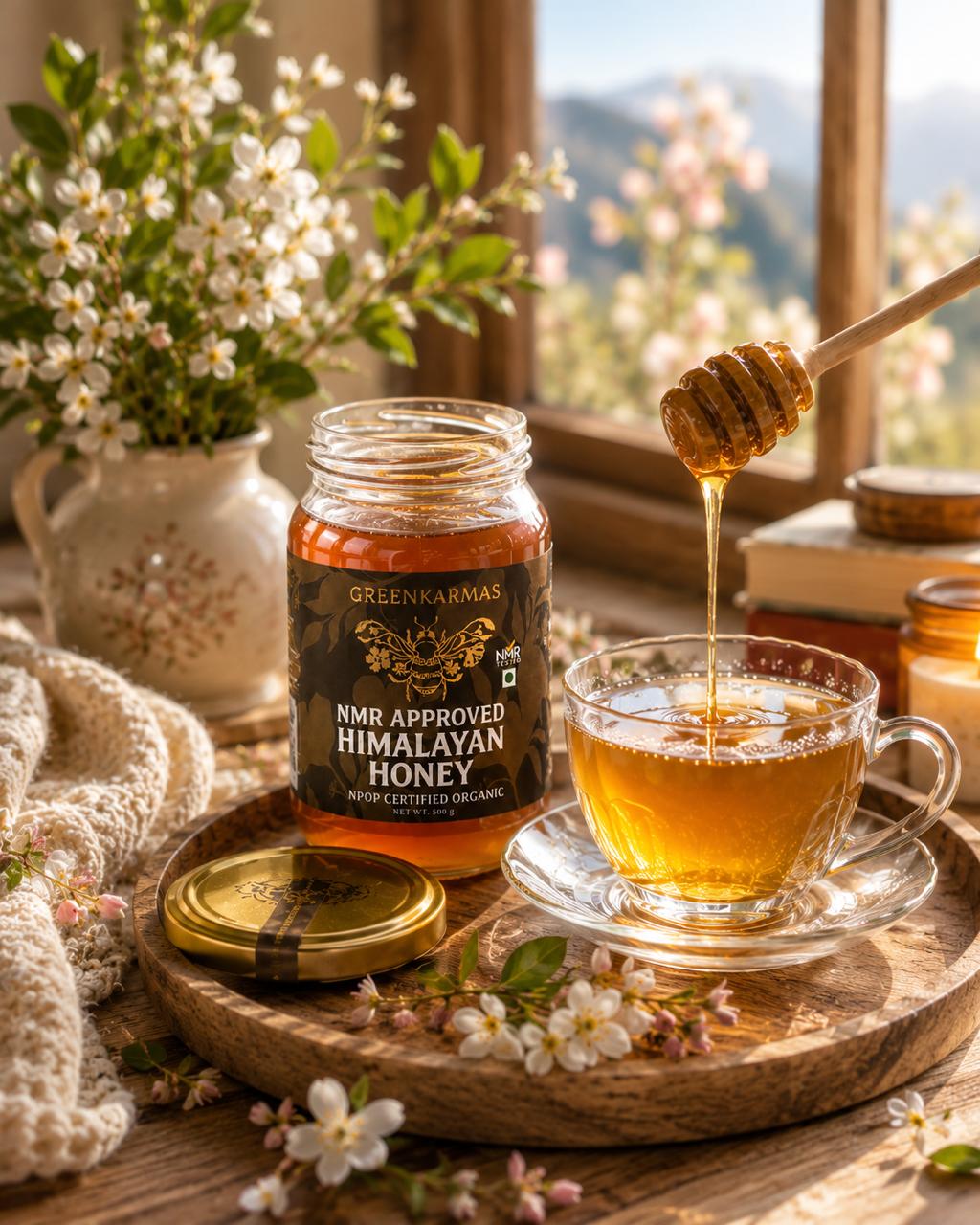 A jar of Greenkarmas NMR Approved Himalayan Honey sits on a wooden tray next to a glass cup filled with honey and a honey dipper, with a window, flowers, and a cozy interior background.